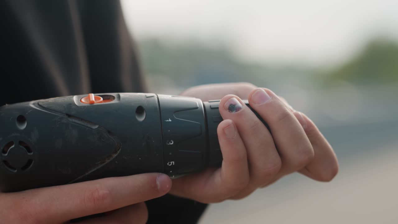 Closeup hands holding cordless drill preparing bit, inspecting chuck and aligning screw, outdoor rooftop background, soft daylight, casual clothing, steady grip, subtle nail polish, shallow depth