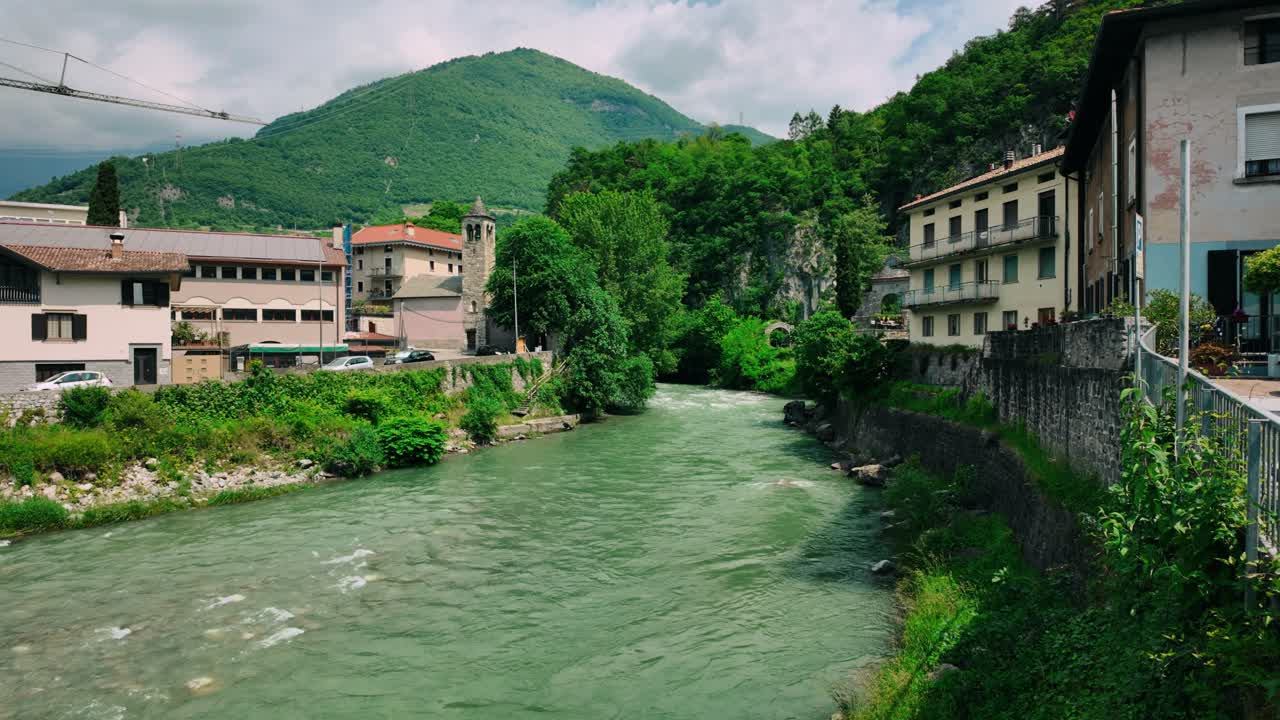 Il fiume Oglio scorre tra tradizionali case in pietra e il campanile a Cividate Camuno. Scena pittoresca del villaggio con una cima montuosa sullo sfondo e una vegetazione lussureggiante lungo l'acqua impetuosa.