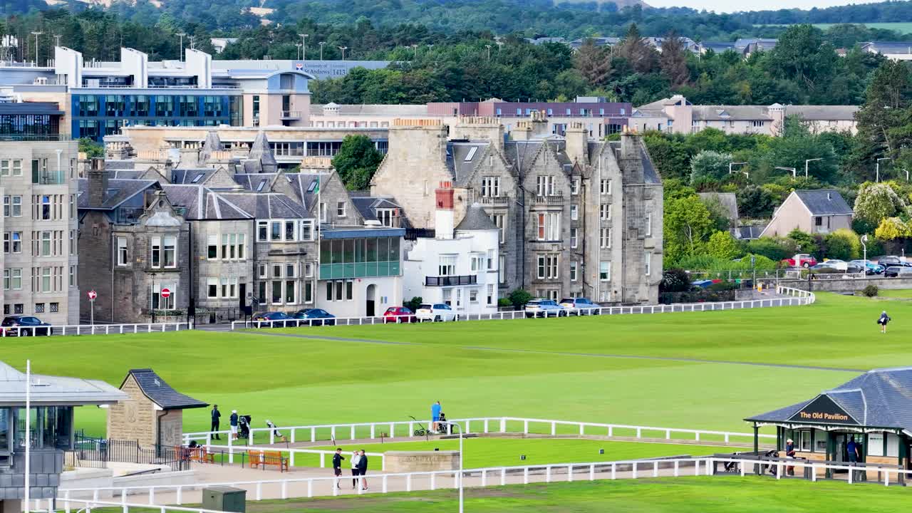 Wide shot of golfers and visitors walking on green, overcast daylight, historic buildings in background