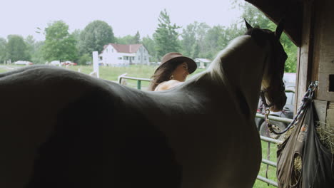 Attractive-young-rider-in-cowboy-hat-smiles-as-she-grooms-her-adult-pinto-horse-in-a-barn