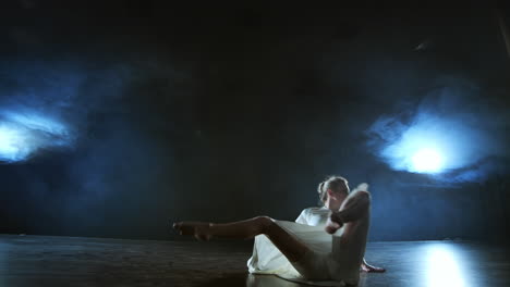 a young female ballerina barefoot jumps on stage and moves in slow motion in a loose white dress.