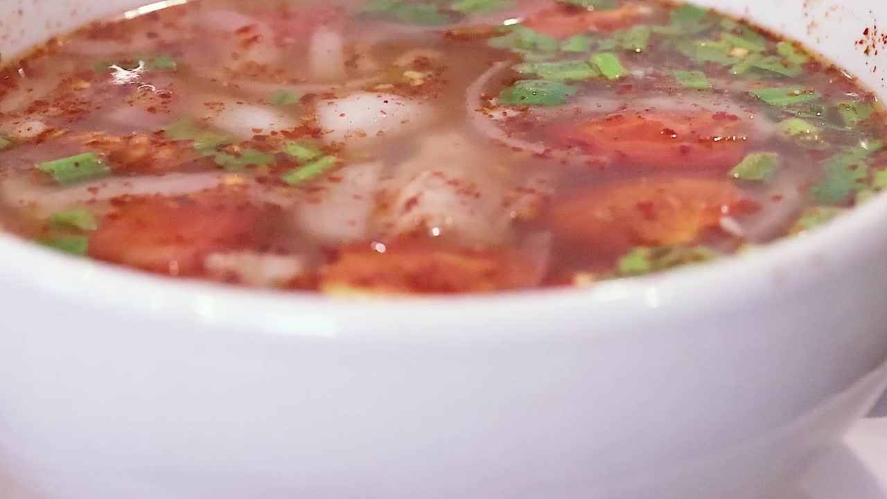 A close-up view of a steaming spicy soup garnished with fresh herbs and vegetables.