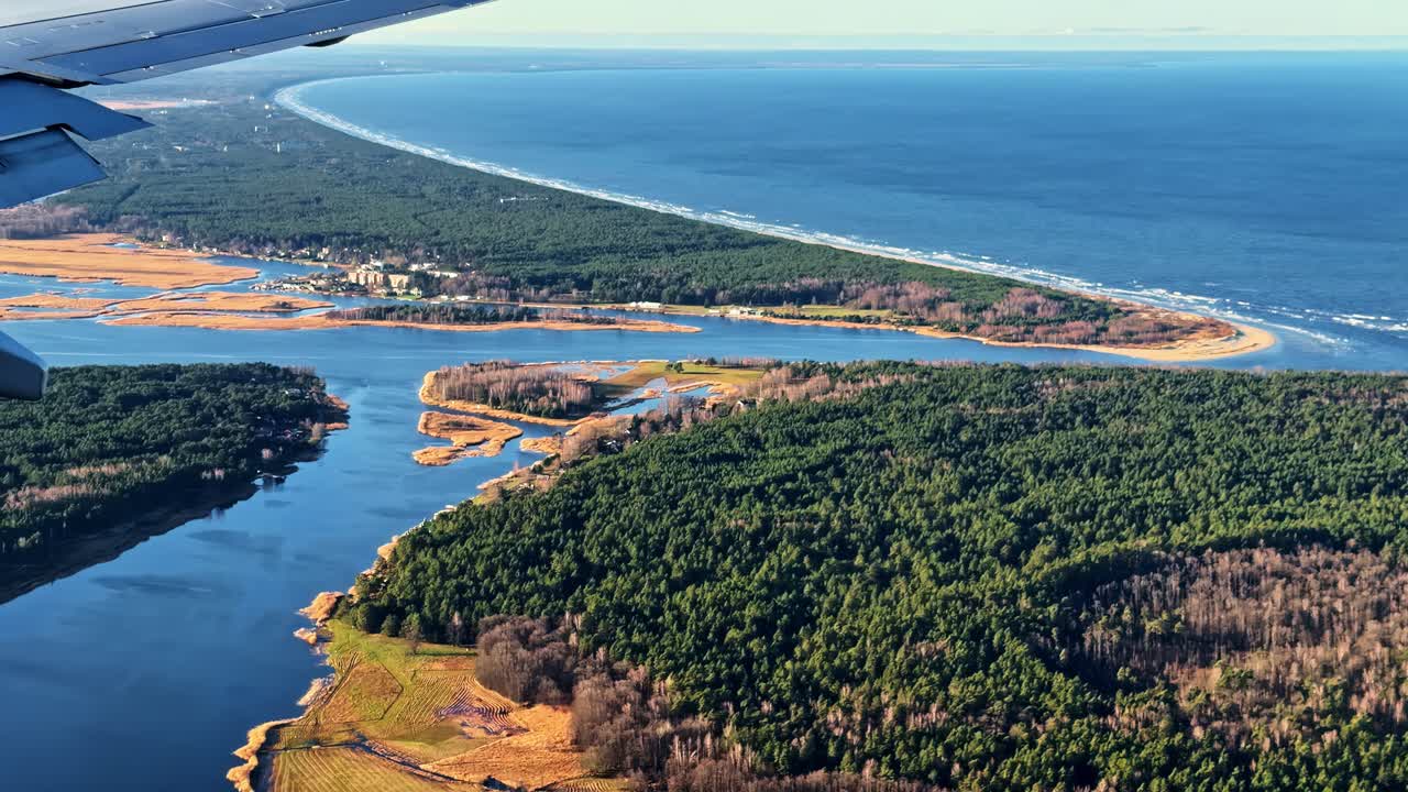 Letecký pohled na lotyšské pobřeží a říční deltu u Rigy, pořízený z okna letadla, zachycující lesní krajinu, klikaté vodní cesty a horizont Baltského moře.