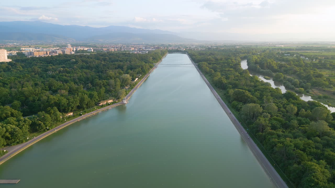 Distant aerial over Plovdiv’s rowing canal at sunset, spring greenery, soft clouds, calm water, and the Rhodope Mountains on the horizon