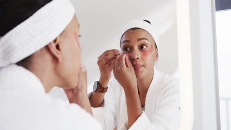 Focused-biracial-woman-applying-under-eye-masks-looking-in-mirror-in-sunny-bathroom,-slow-motion