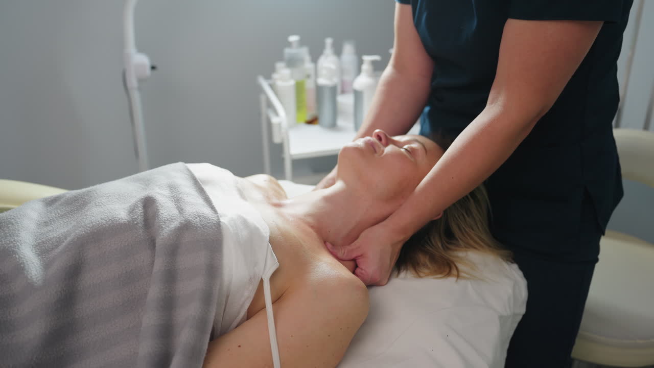 Close up of beauty specialist smiling as gliding fingertips along side of client neck to shoulder during relaxing spa treatment in serene clinic environment with soft linens and gentle lighting