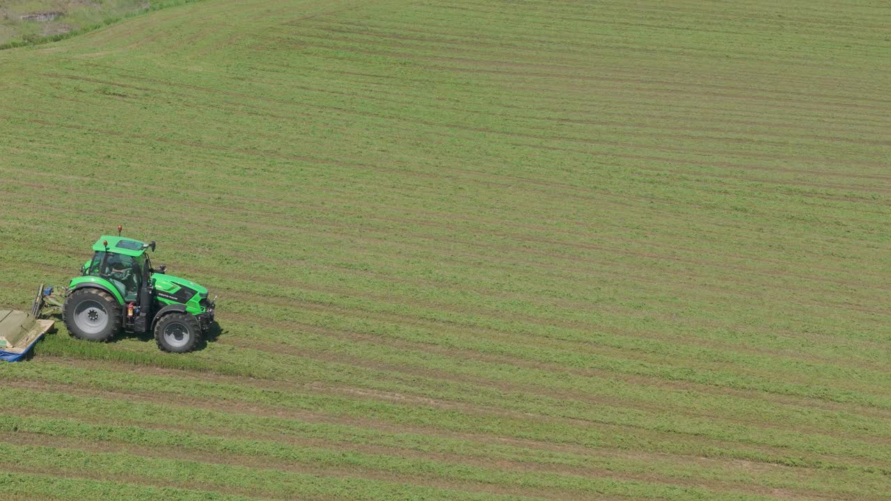 Green tractor cutting grass in large field, creating neat parallel rows, drone shot.