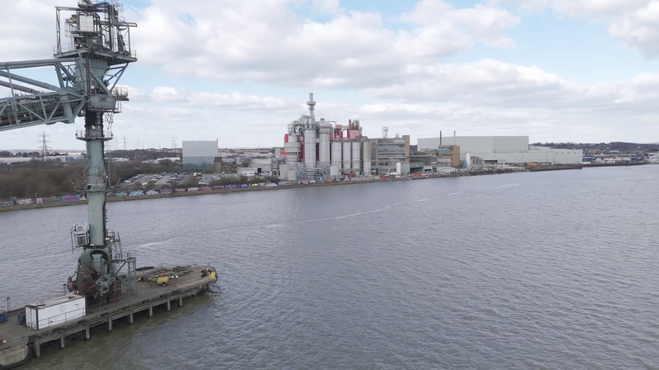 Aerial sweep of riverside industrial zone featuring warehouses, cranes and loading arms projecting over calm water, reflecting large-scale logistics and manufacturing operations