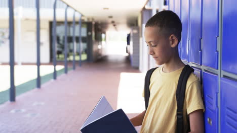 biracial boy with backpack stands by blue lockers, reading a book, copy space