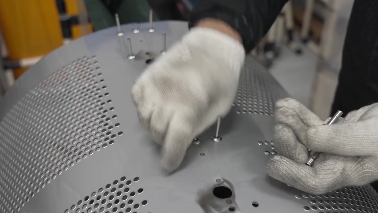 Worker inserting metal rivets into perforated stainless steel washing machine drum bottom plate at workbench