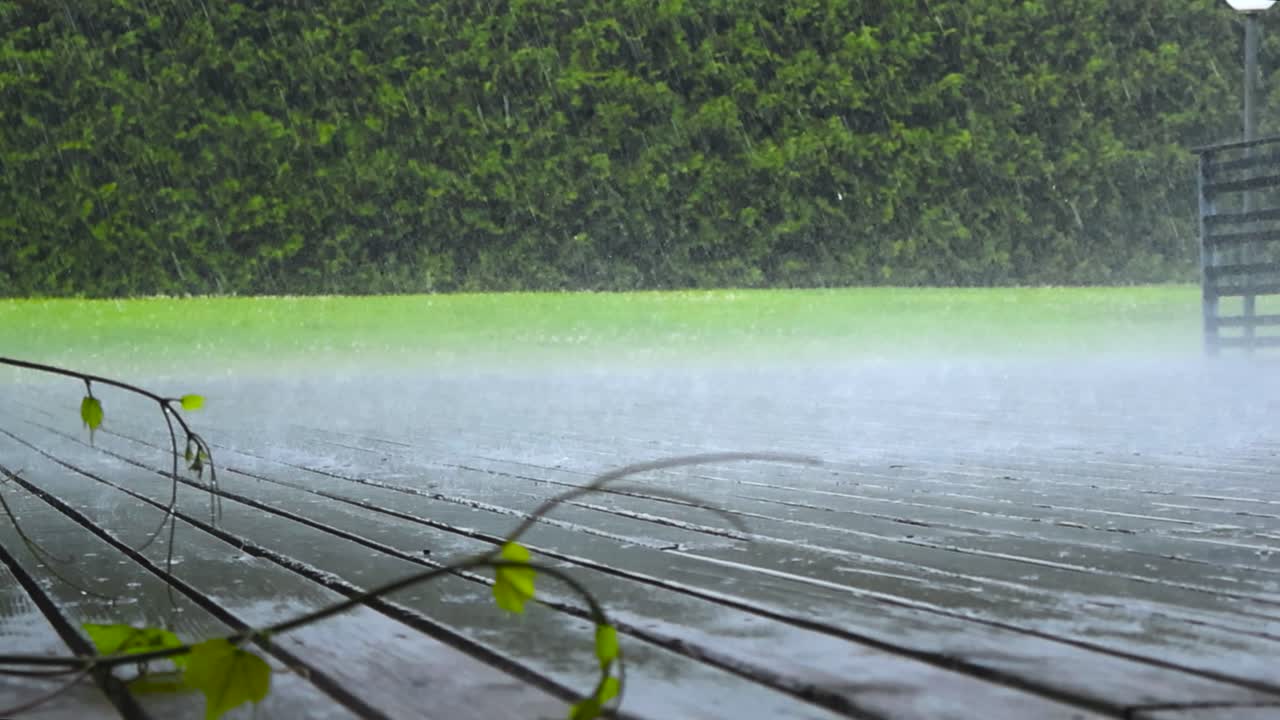 Low angle video of heavy rainfall and hale falling on a wooden deck or a terrace during a summer or autumn day with ice pieces visible bouncing and green cut and mowed lawn grass in the wet background