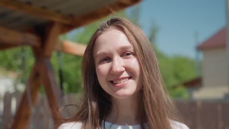 cheerful young girl with long straight hair smiling gently under bright sunlight, standing outdoors near wooden structure with green trees and wooden fence in background on peaceful sunny day