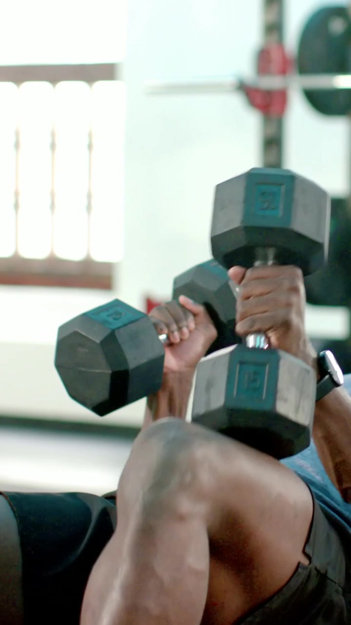 Vertical video: Knee bracing, gripping dumbbells, man in gray tee pressing chest on bench, tracker