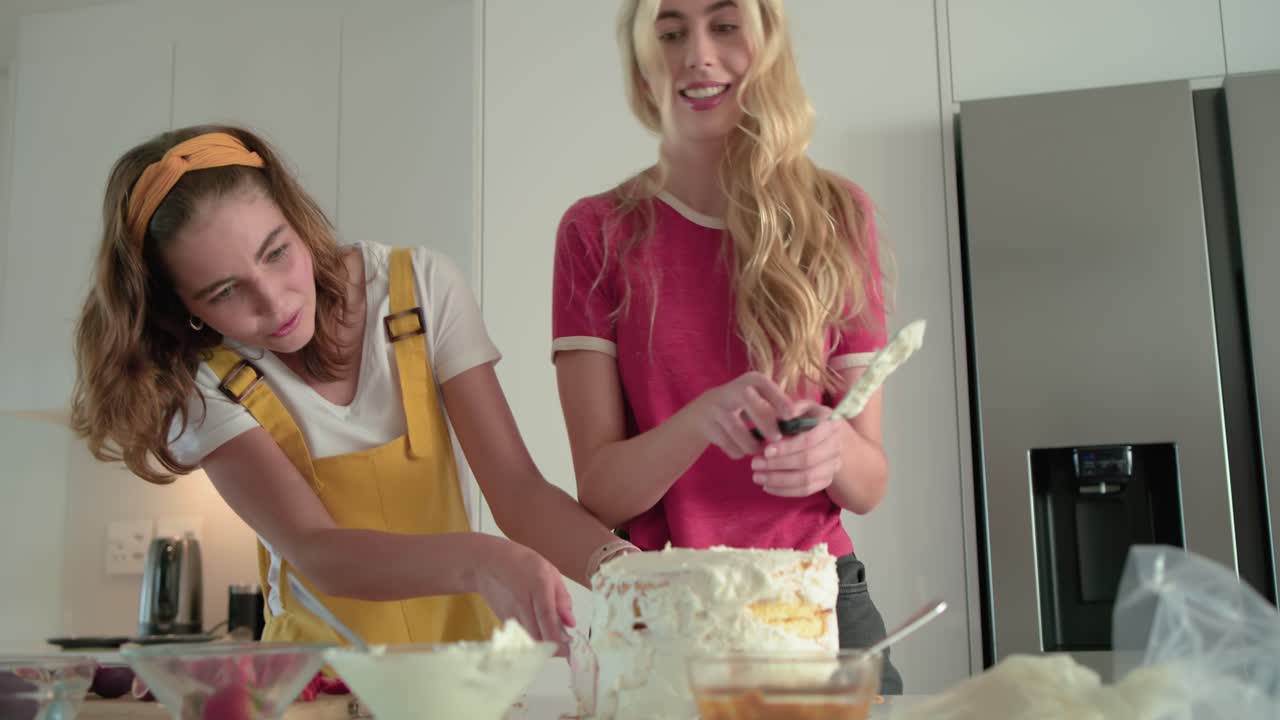 Seeing frosting and fruit bowls, two women smoothing icing with spatula on cake at kitchen counter