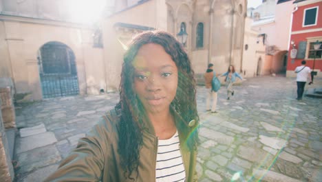 mujer afroamericana con cabello rizado sosteniendo un teléfono inteligente y saludando a la cámara mientras sonríe en la calle