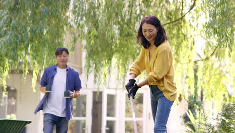 mature asian woman raking and tidying leaves in garden as husband brings her hot drink in cup - shot in slow motion