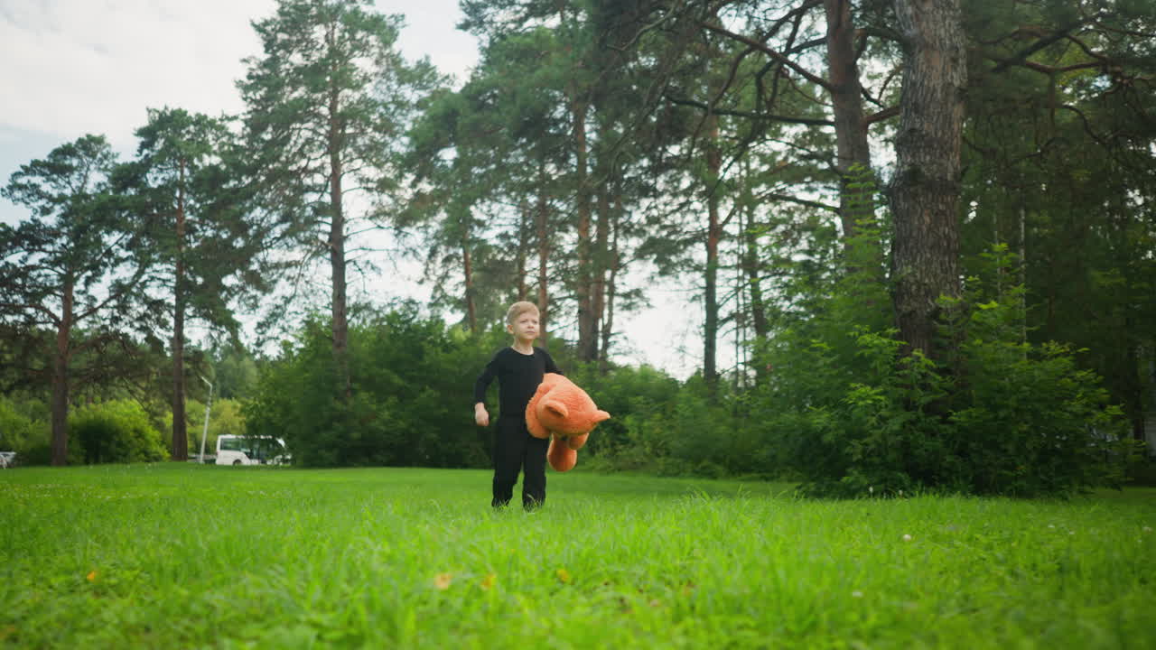 Young boy in black outfit carrying large teddy bear while walking through lush green open park surrounded by tall trees, looking thoughtful with soft natural light and relaxed atmosphere