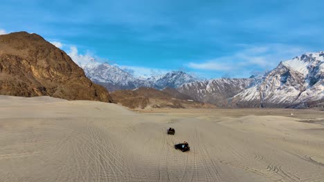 aerial view of sarfaranga cold desert shigar with suvs driving over sand dune with snow covered mountains in background. orbit motion shot