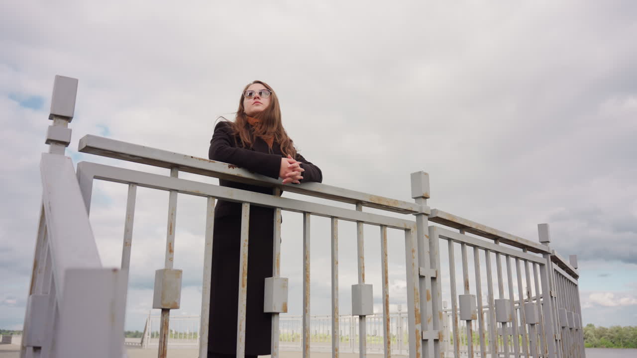 Tall lady in long dark winter coat stands under hazy sky near iron safety railings, viewed from low angle, expressing calm posture, elegance, and confidence in peaceful outdoor urban environment