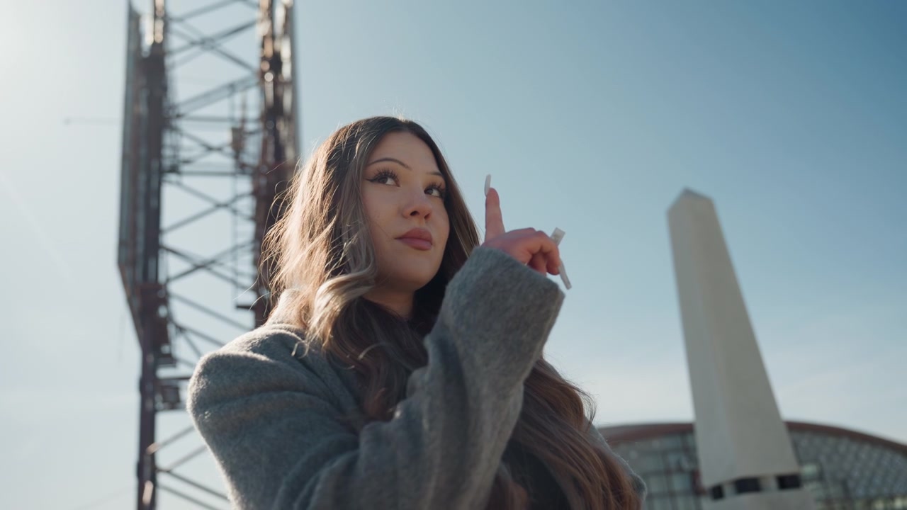 Gesto giocoso sul tetto di una donna asiatica, bella guida che indica verso l'alto con un sorriso pensieroso e un bordo di controluce caldo, torre reticolare industriale e obelisco che incorniciano la scena, cappotto accogliente e capelli lunghi, speranzosa.