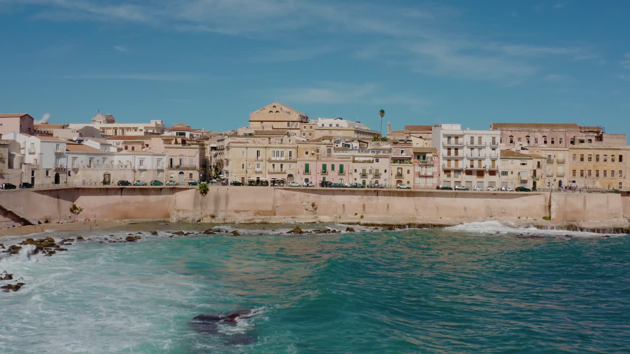 Aerial drone shot of Ortigia island in Syracuse. Flying towards the coast with large waves crashing on the shore. Medieval natural fortress around the city. Historic Italian town in Sicily.