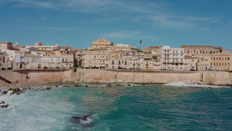 aerial drone shot of ortigia island in syracuse. flying towards the coast with large waves crashing on the shore. medieval natural fortress around the city. historic italian town in sicily.