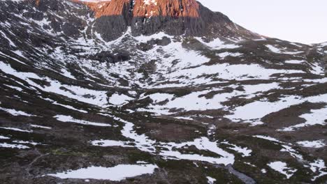 Aerial-drone-footage-revealing-Hutchison-Memorial-Bothy-and-warm-sunrise-over-a-dramatic-vertical-cliff-rising-out-of-the-snow-and-moorland-in-winter