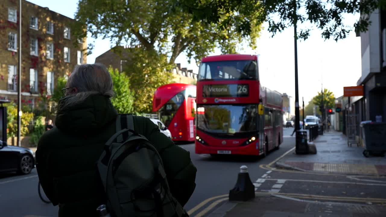 Man with glasses waits for red double-decker bus route 26 to Victoria in London street scene