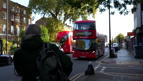 man with glasses waits for red double-decker bus route 26 to victoria in london street scene