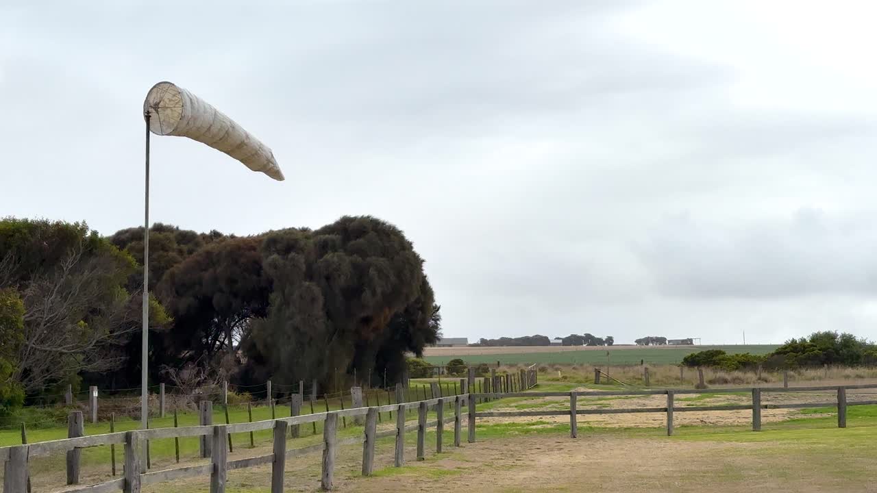 A windsock flutters in a breezy coastal landscape, surrounded by greenery and overcast skies, captured in a serene setting