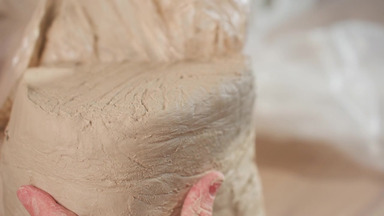Caucasian potter inspects wedged clay up close, hands smoothing cylinder and checking moisture, fingertips marking surface, bench light revealing texture, quiet focused studio moment before throwing
