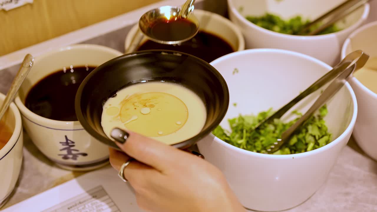 Hand pours spicy chili oil onto sesame sauce bowl at self-serve hotpot condiment station