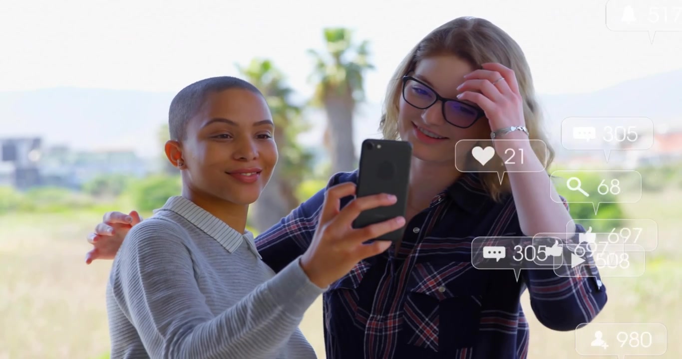 Two friends raising smartphone, framing selfie, generating floating social icons for social sharing