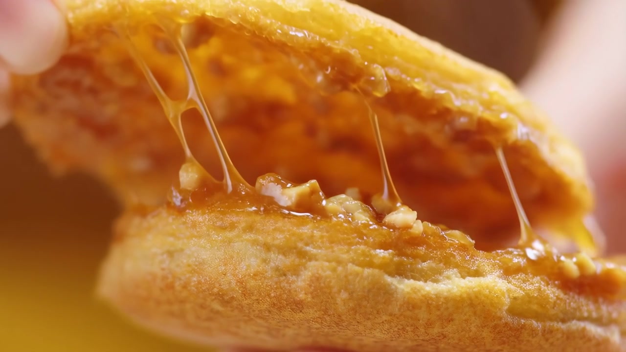 Dedos delicados abrem uma massa de sobremesa macia, espalhando fios de caramelo para fora, enquanto pedaços de nozes e recheio âmbar brilhante aparecem através da crosta folhada, e a luz quente realça a textura pegajosa e a cor rica.