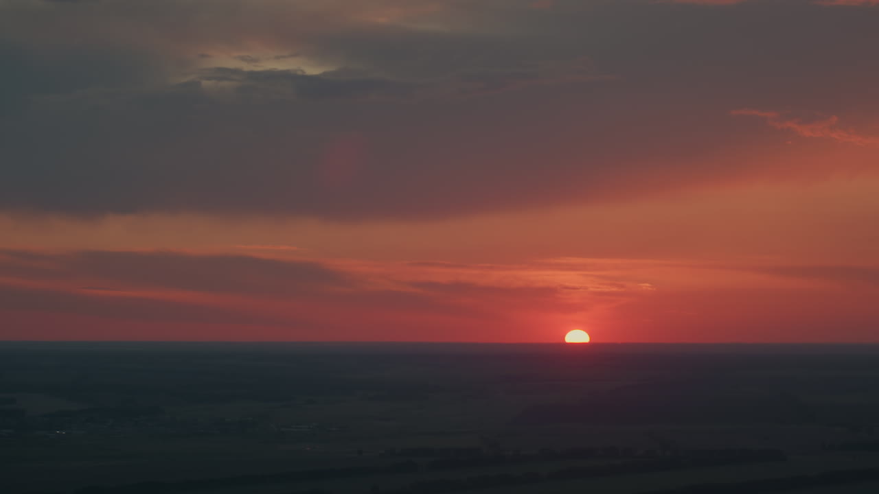 Towering dusk clouds over vast farmland horizon illuminated by vibrant red and orange sunset glow with subtle lens flare and sky layering