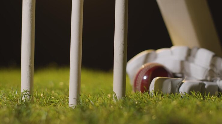 Natura morta di cricket con primo piano di mazza, palla e guanti adagiati sull'erba dietro i paletti