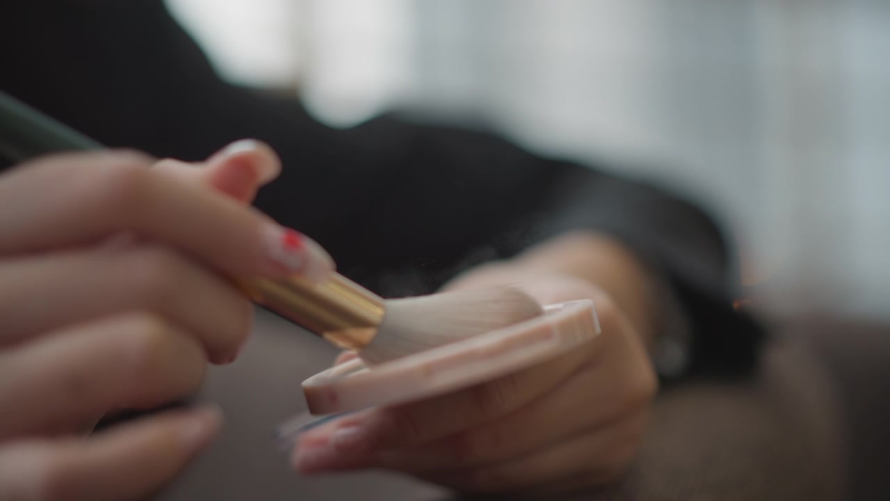 Close view of hand sweeping fluffy makeup brush across loose light beige face powder inside round compact, showing gentle cosmetic application, soft bristles, disturbed texture, and elegant grooming