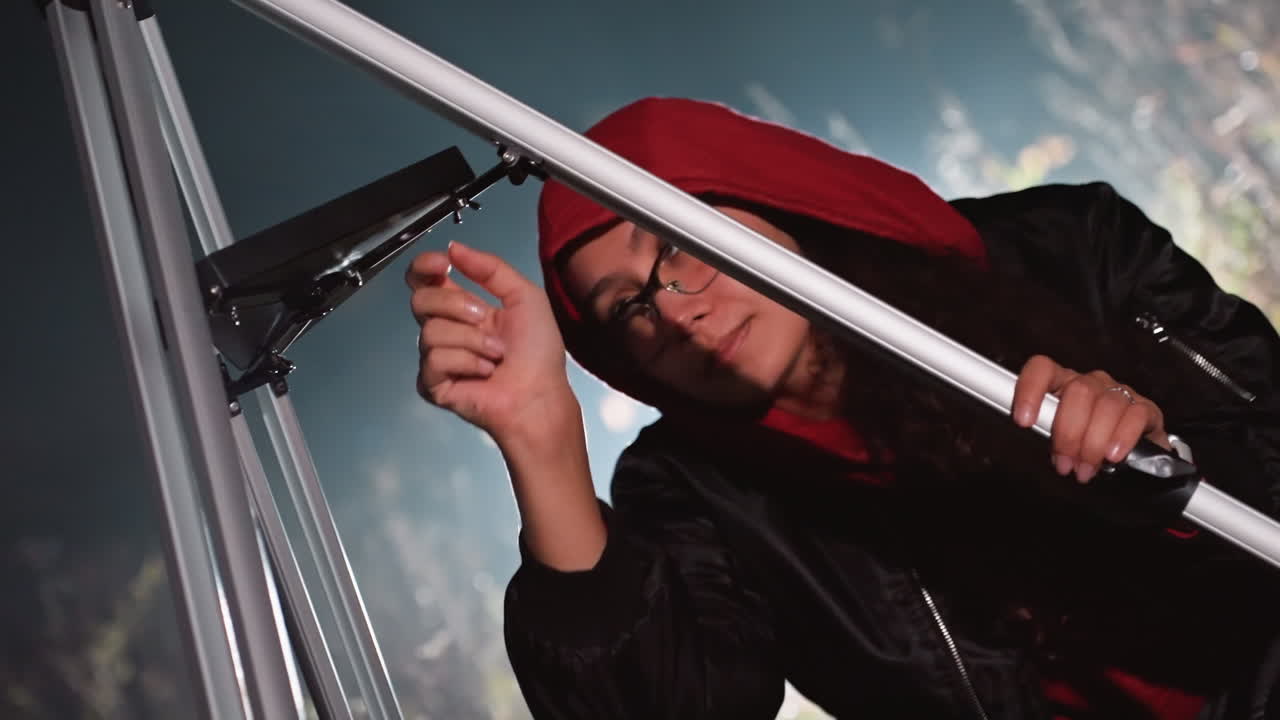 Astronomo che regola il telescopio sotto il cielo notturno, indossa una felpa con cappuccio rossa e occhiali, calibra il supporto in metallo sull'osservatorio sul tetto con bokeh di luci della città, mano attenta che regola la manopola di messa a fuoco, metodico