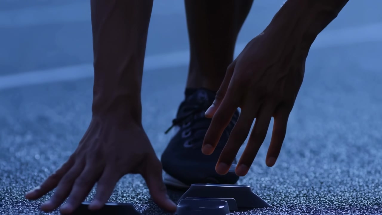 Primo piano a livello del suolo che mostra le mani e la scarpa di un corridore che premono sui blocchi di partenza in plastica su una pista di gomma testurizzata, con illuminazione blu fredda, tensione, concentrazione e preparazione alla gara, con corridore, mano.