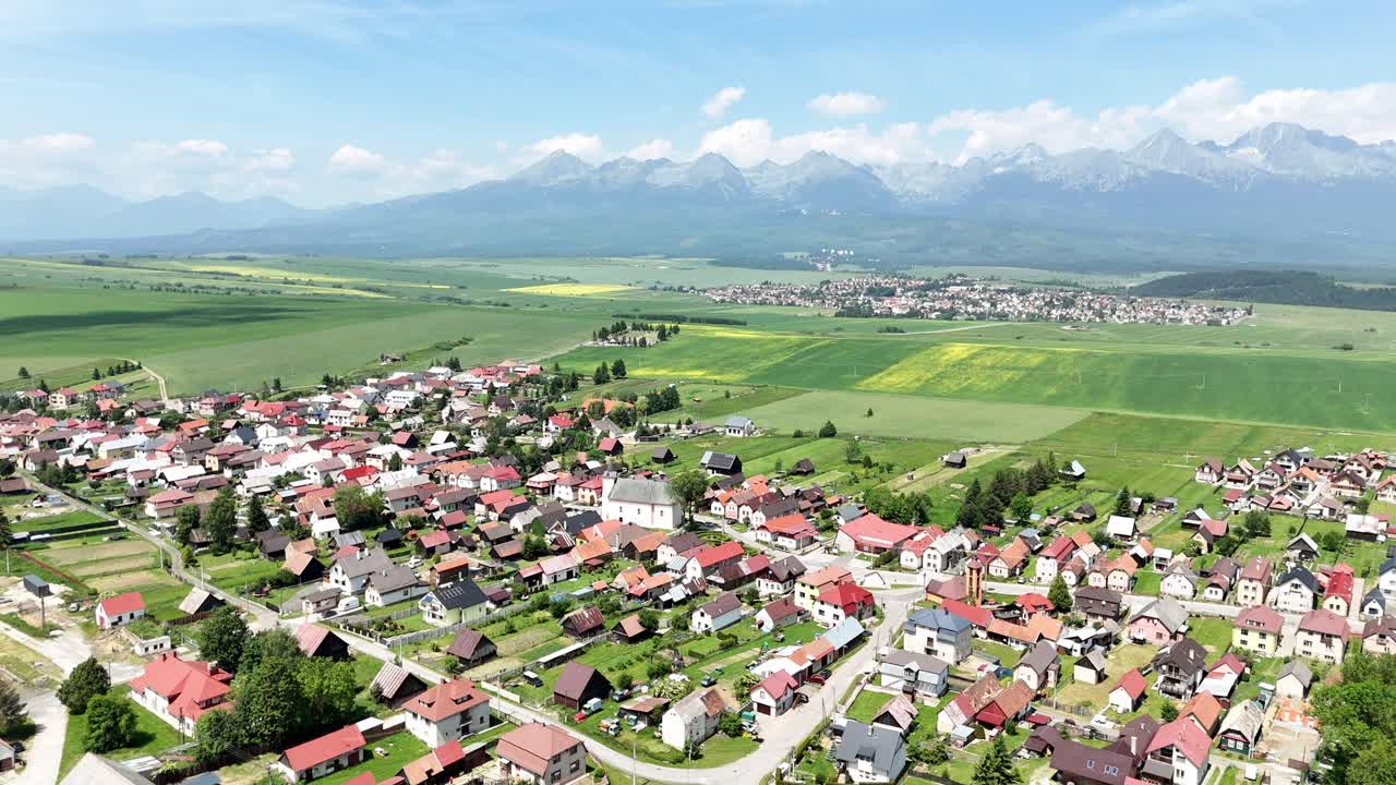 The drone slowly descends over the Slovak countryside, flying toward a small church surrounded by green fields, with the High Tatras mountains visible in the distance under a bright blue sky
