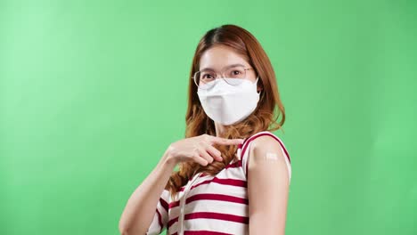 Young-Asian-woman-pointing-to-her-arm-after-being-vaccinated-against-covid-19-in-studio-with-chroma-key-background