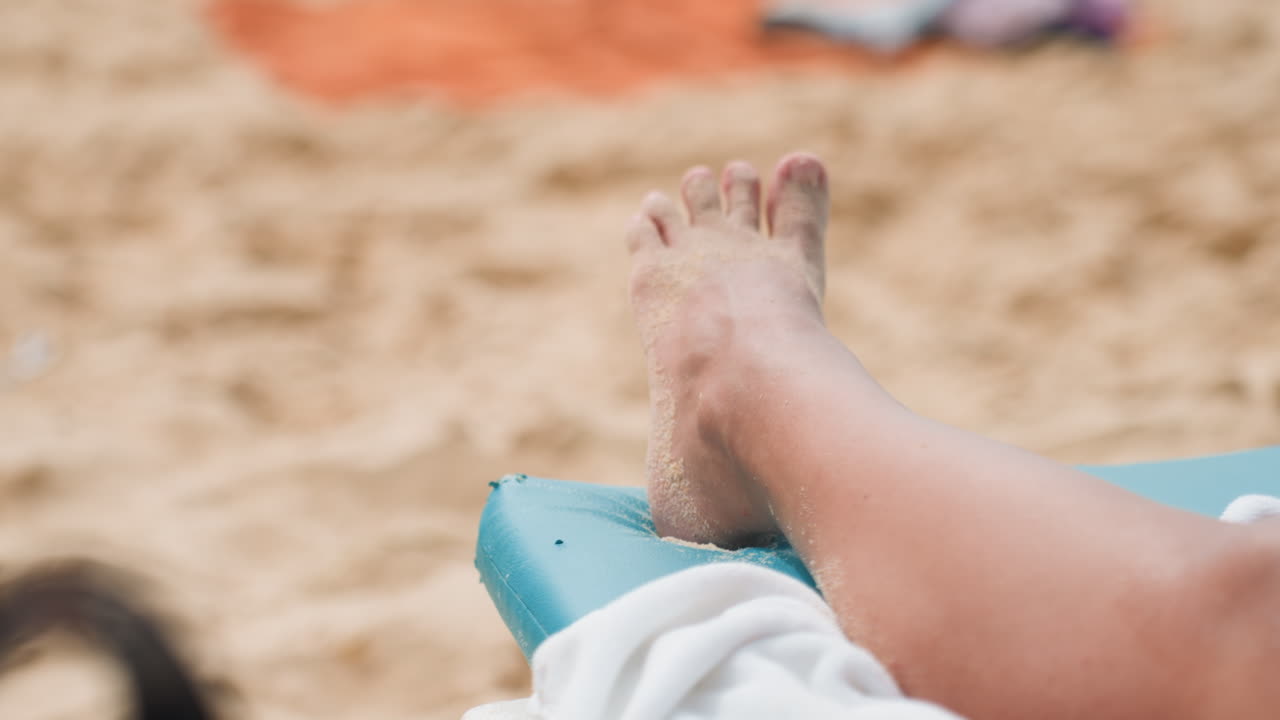 A piedi nudi su una spiaggia sabbiosa dell'isola, un vacanziere sdraiato su una sdraio turchese, primo piano di un piede coperto di sabbia appoggiato su un asciugamano, luce solare calda e brezza leggera, atmosfera tropicale rilassata per un video al rallentatore.