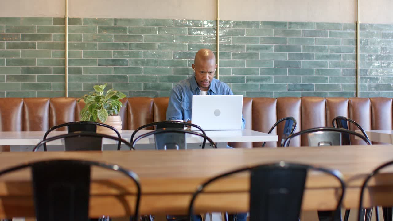 African man in blue shirt working at cafe, responding to camera focus, typing on silver laptop