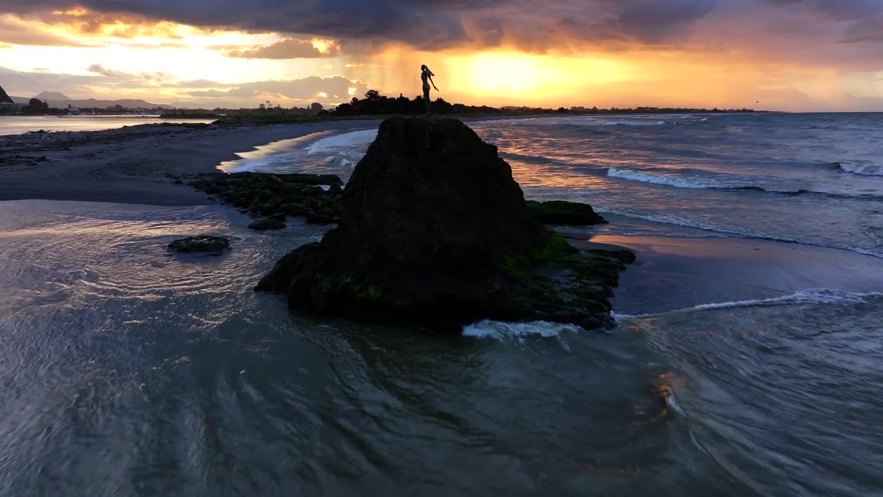 Sunset waves crash against a rocky outcrop, spray rising around a lone statue silhouetted in gold—still and solemn amid the ocean’s restless glow.