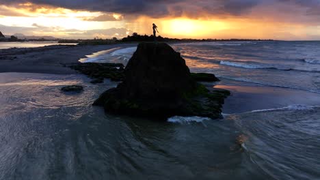 sunset waves crash against a rocky outcrop, spray rising around a lone statue silhouetted in gold—still and solemn amid the ocean’s restless glow.