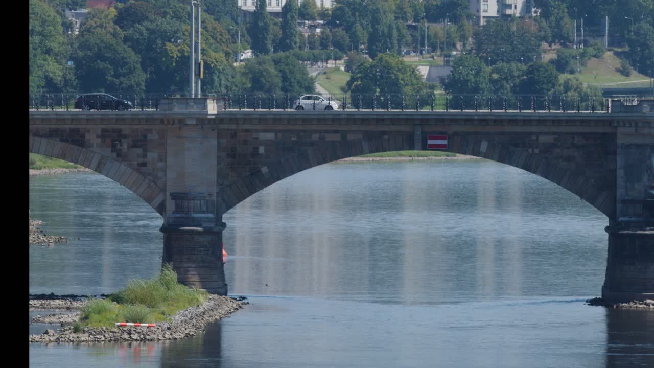 Daytime telephoto sequence of cars and a red bus traveling across a stone arch bridge in Dresden, Germany, with calm river and urban greenery