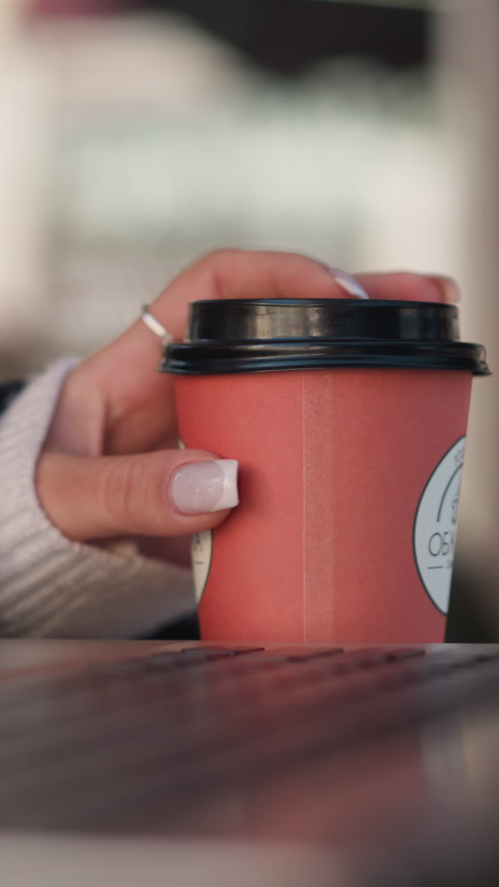 primer plano de la mano de la señora girando suavemente la taza de café cerca de la computadora portátil mientras usa el ratón, resaltando sus dedos manicurados, con un fondo borroso del espacio de trabajo y el entorno moderno