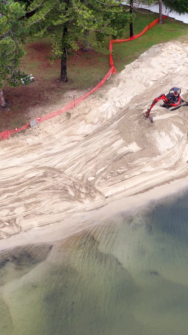 Vista Aerea di un Escavatore Rosso che Sposta Sabbia per il Ripristino della Spiaggia Lungo un Canale sul Lungomare