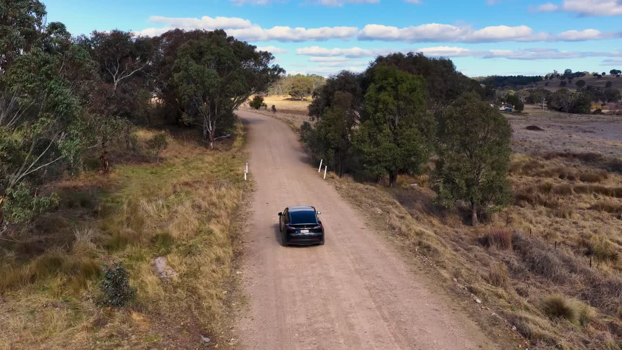 A black electric vehicle travels down a remote dirt road in rural Queensland, Australia, captured by a smooth aerial drone shot in daylight