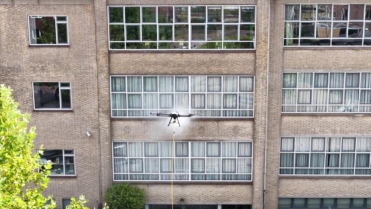 Tecnologia avanzata di droni commerciali che utilizza acqua ad alta pressione per un servizio automatizzato di pulizia finestre sulla facciata di un grande edificio per uffici in mattoni, mostrando l'innovazione futura nella manutenzione.